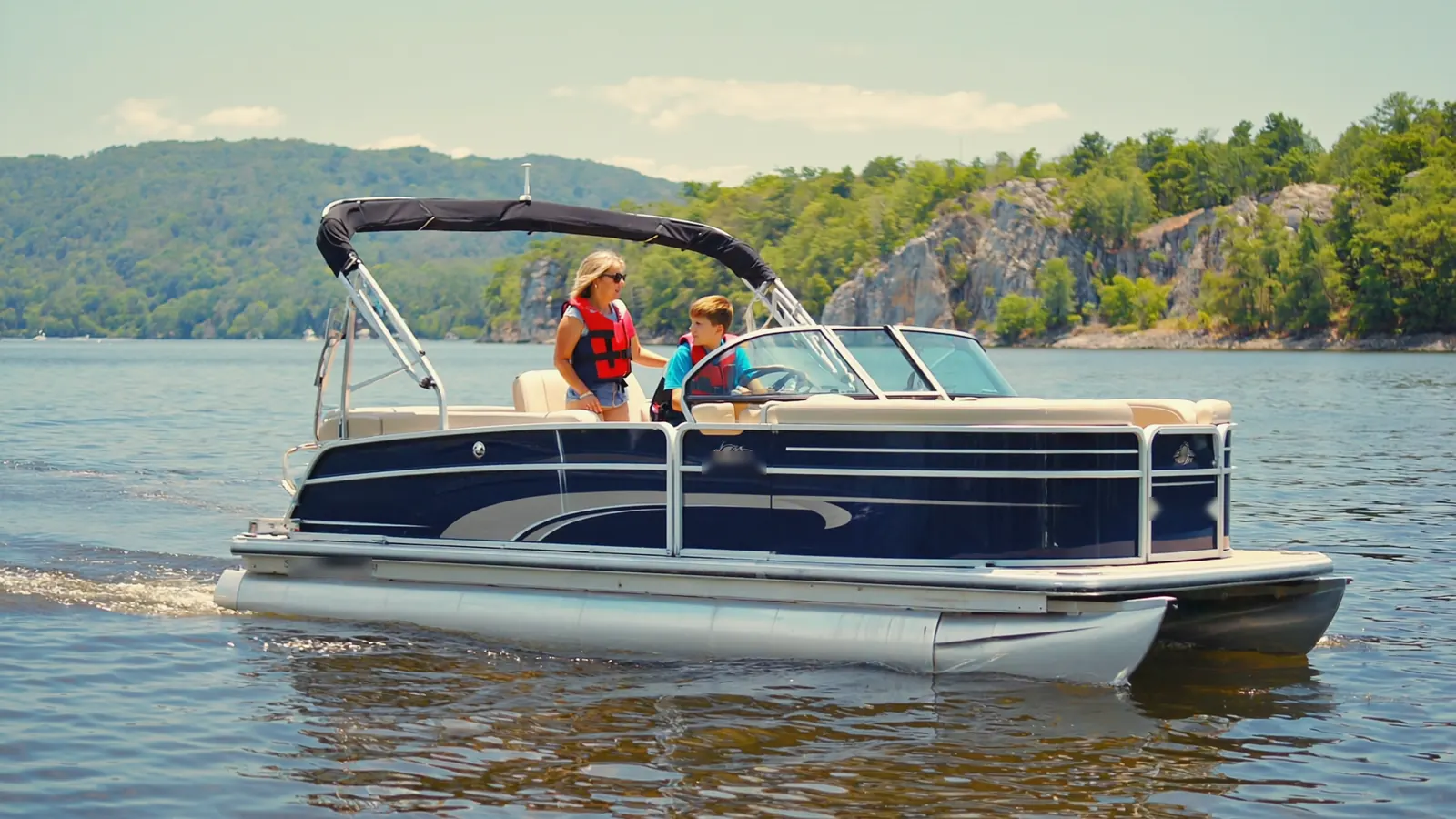 A mother stands beside her teenage son at the helm of a pontoon boat on a bright day, both wearing life jackets