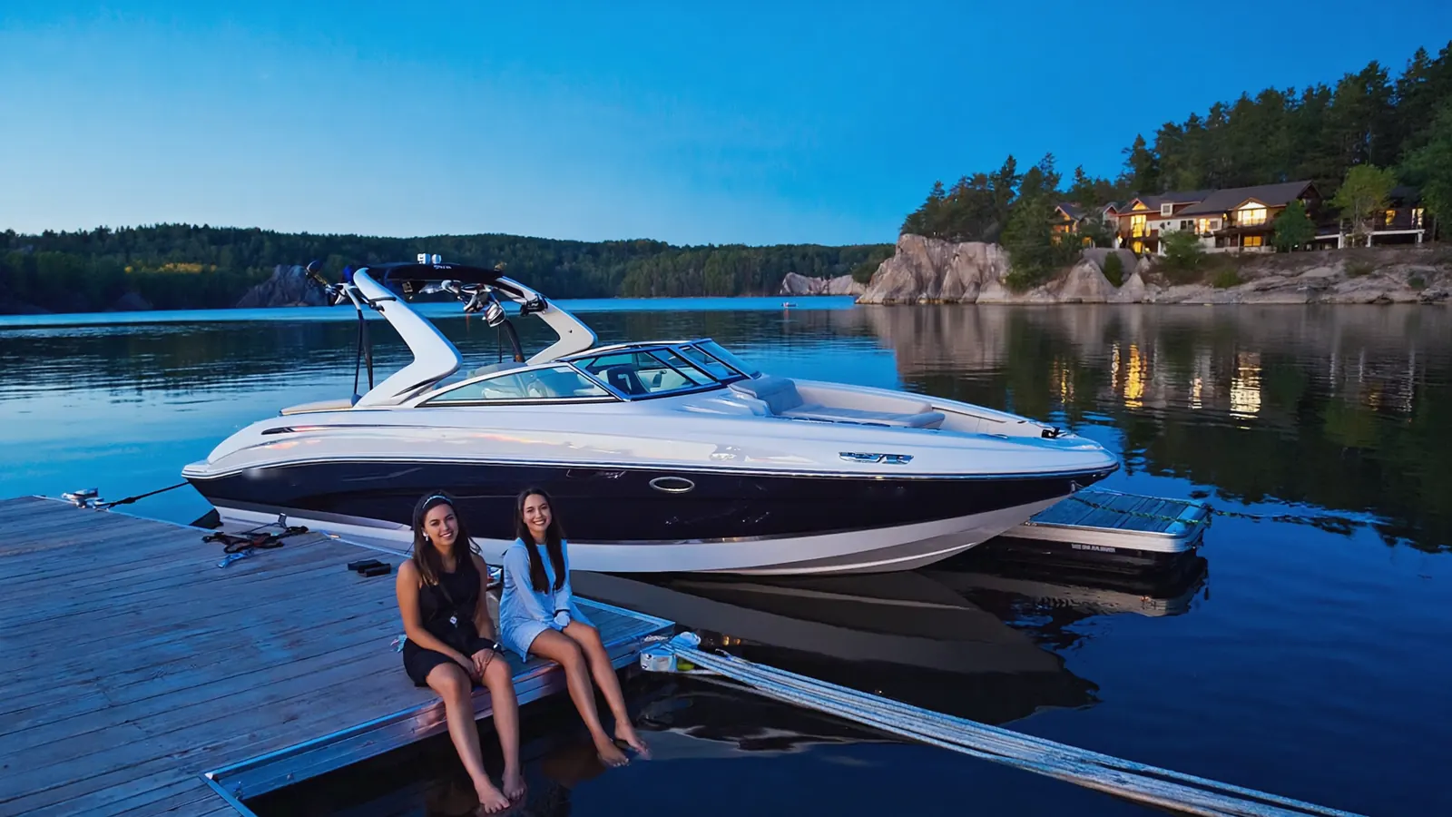 Two friends sitting on a dock beside a moored bowrider at blue hour with lake-house lights on the water