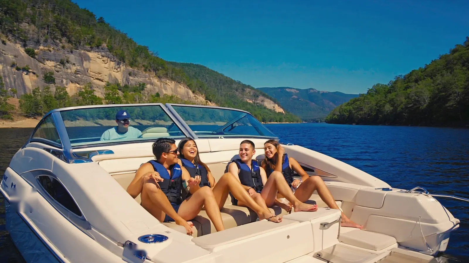 Four friends in life jackets laughing in the cockpit of a bowrider while the driver sits at the helm, Tennessee bluffs behind