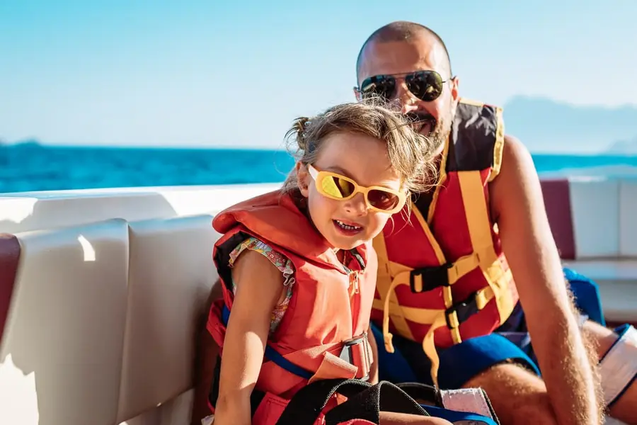 A father and young daughter wearing life jackets on a boat in bright sunlight.